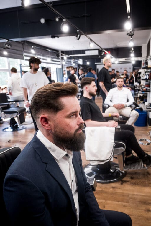 Mężczyzna w garniturze siedzi w zatłoczonym salonie fryzjerskim