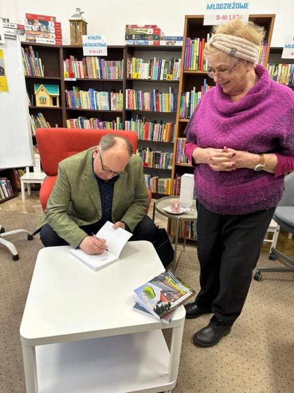 Starszy mężczyzna podpisuje książkę, kobieta obserwuje w bibliotece.