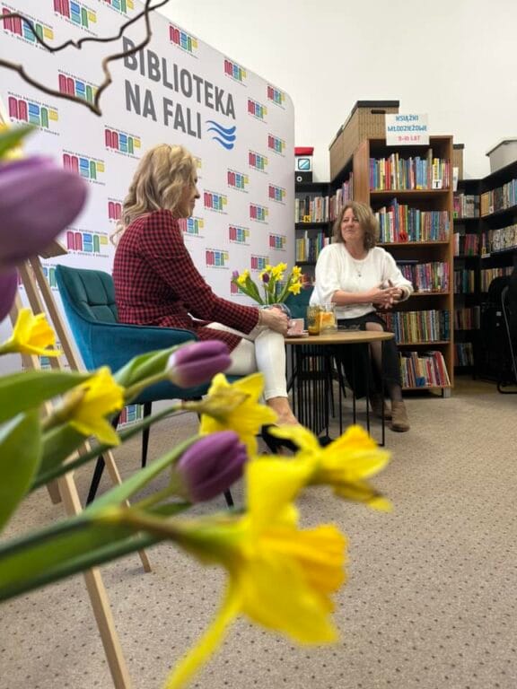 Dwie kobiety rozmawiają w bibliotece z kwiatami na pierwszym planie.