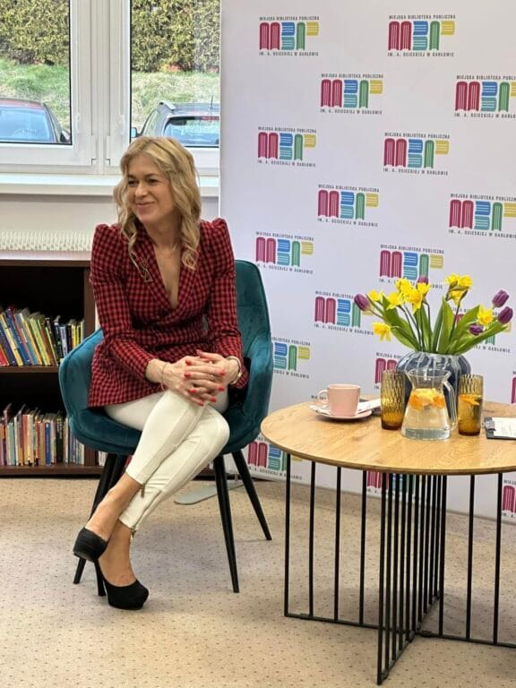 Kobieta siedzi na fotelu w bibliotece, uśmiechnięta