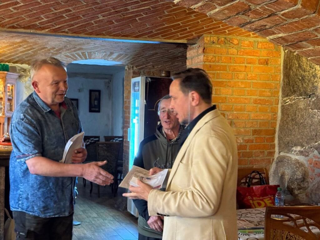 Trzej mężczyźni rozmawiają w rustykalnym wnętrzu.