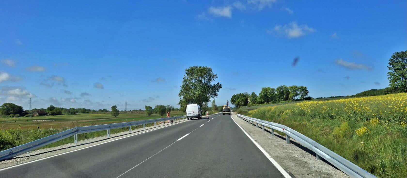 502514338_1103369378483452_4450954959362693864_n Pojazdy na słonecznej drodze z barierkami i polami kwiatów