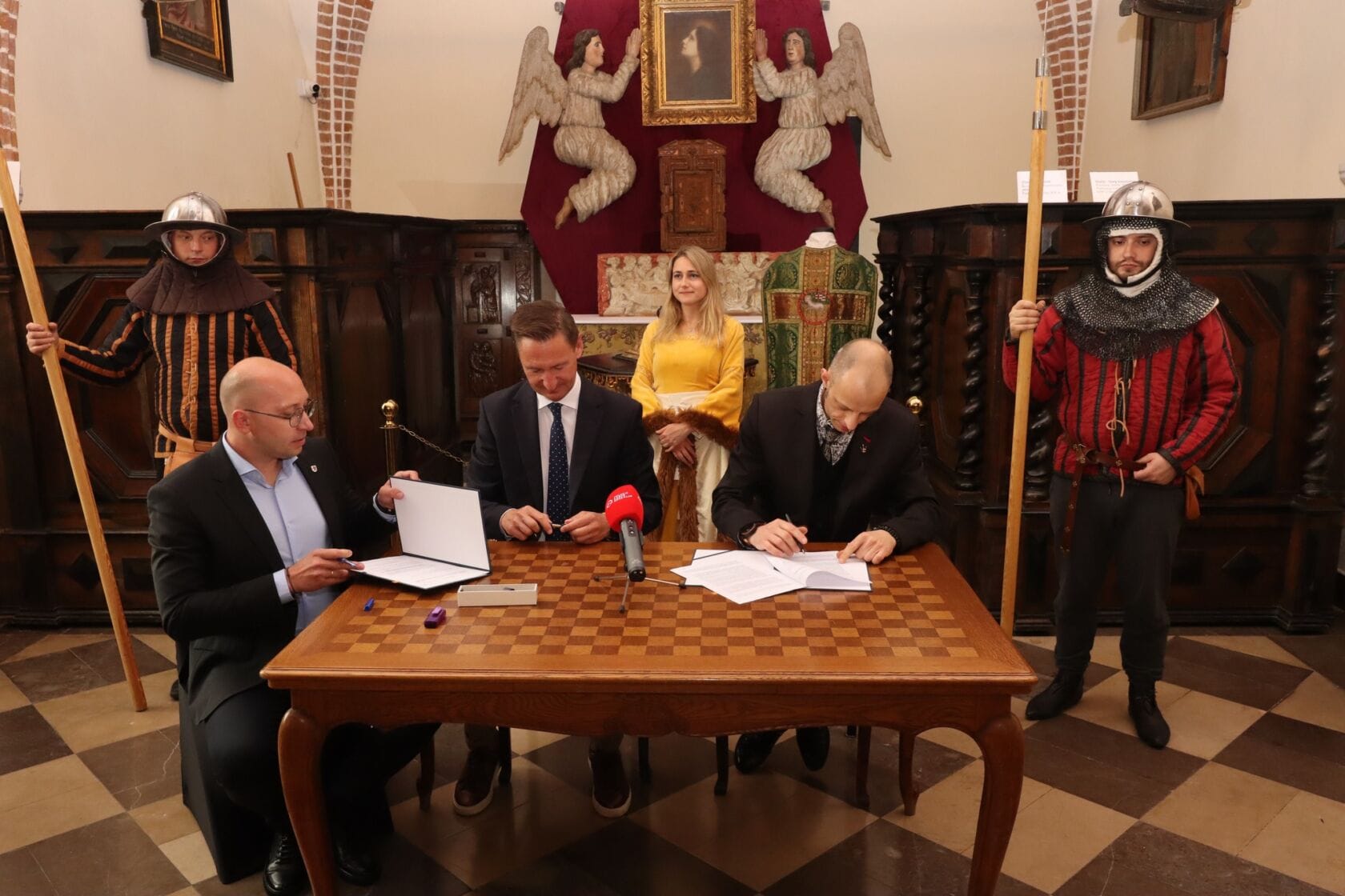 Mężczyźni podpisują dokumenty, kobieta i strażnicy w tle