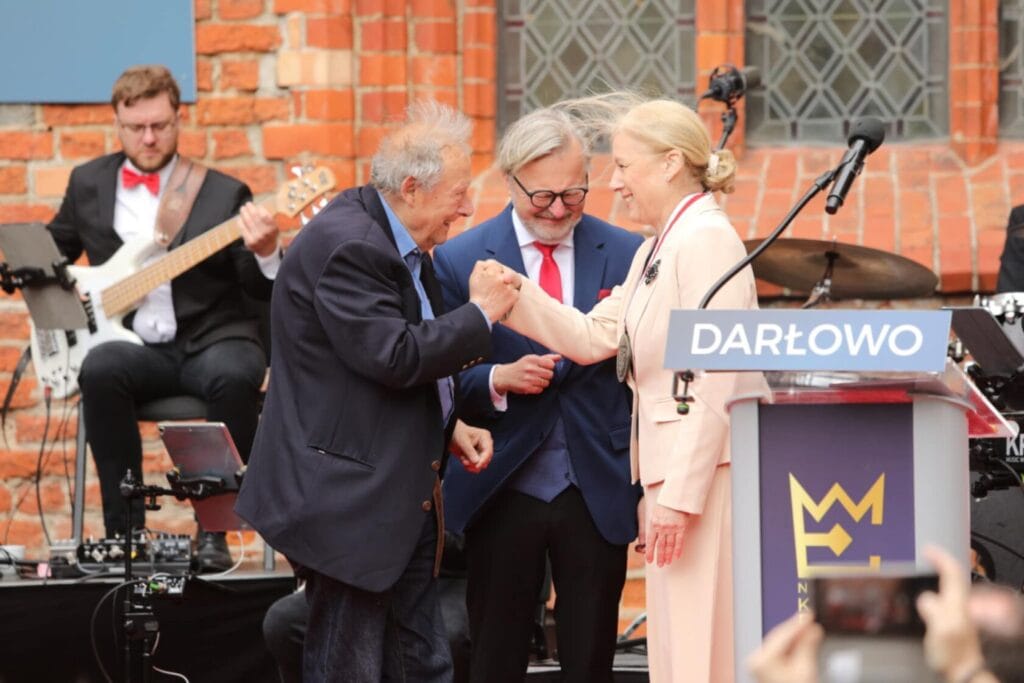 Starszy mężczyzna wita się z kobietą na scenie przy zespole