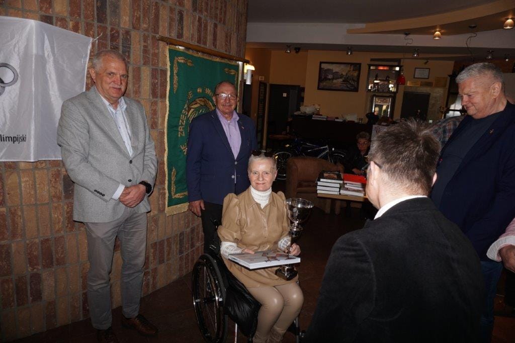 Grupa osób wewnątrz, kobieta na wózku z pucharem