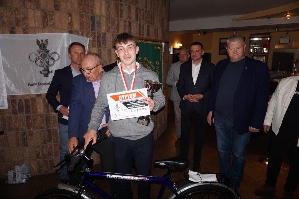 Młody laureat z dyplomem i rowerem na tle flagi.