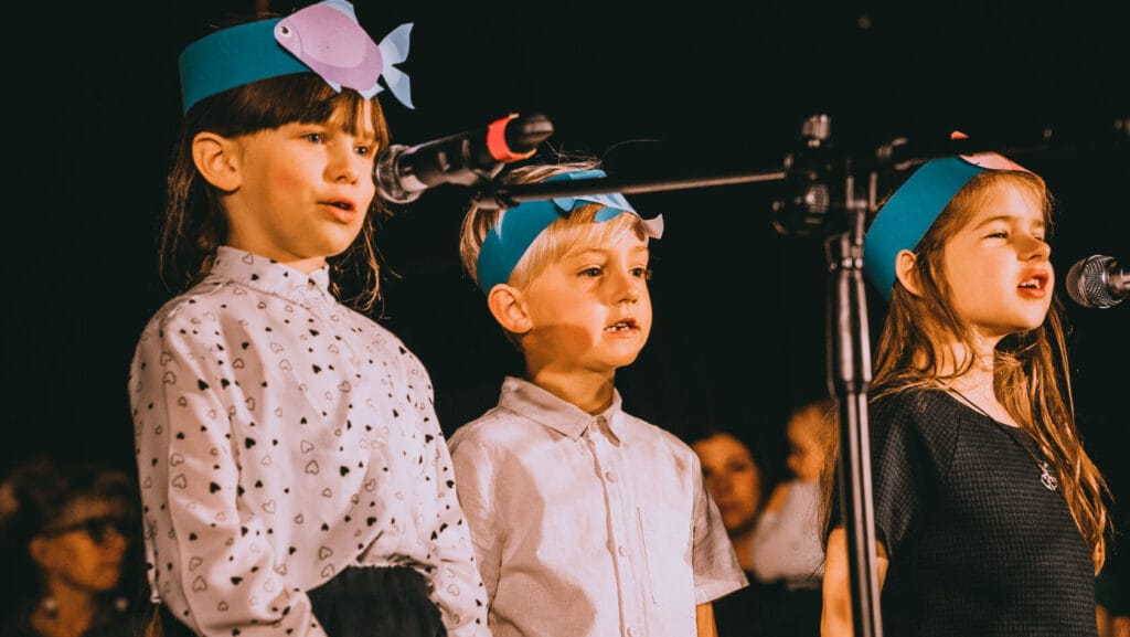 Dzieci śpiewają na szkolnej scenie, noszą kolorowe opaski