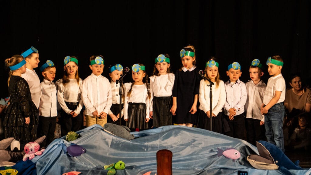Dzieci w opaskach z globusami śpiewają w szkolnym przedstawieniu