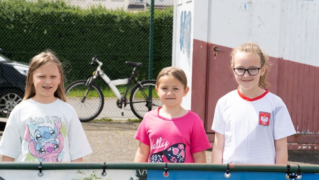 Trzy dziewczynki uśmiechają się stojąc obok ogrodzenia.