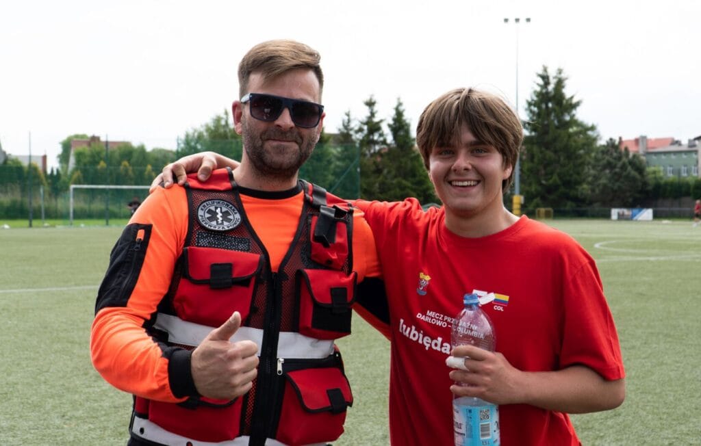 Dwóch uśmiechniętych mężczyzn na boisku, jeden w kamizelce ratunkowej