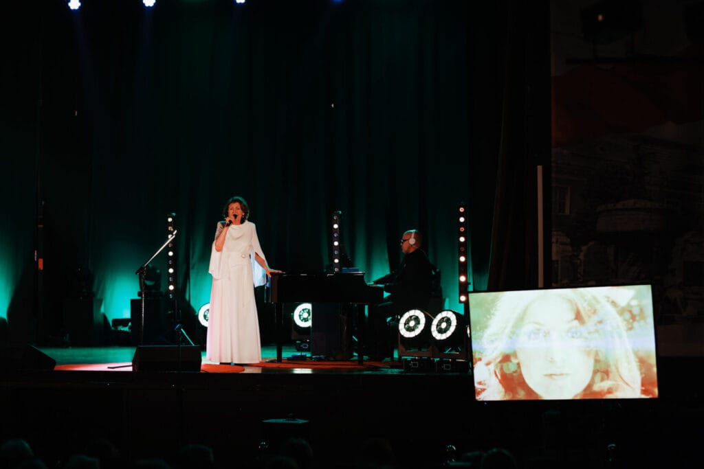 Kobieta śpiewa na scenie z pianistą koncertowym w tle