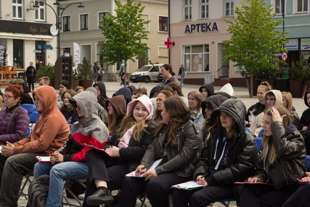 Grupa osób siedzi na krzesłach w miejskiej przestrzeni