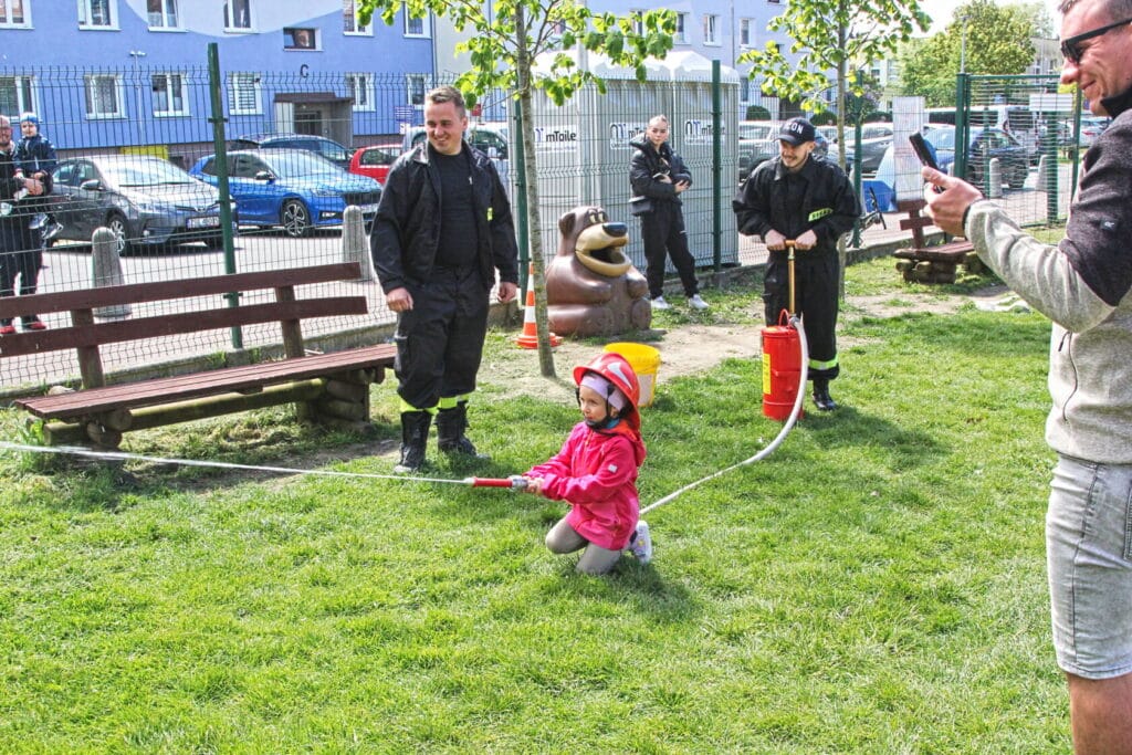 Dziecko w strażackim hełmie trzyma wąż przy fontannie