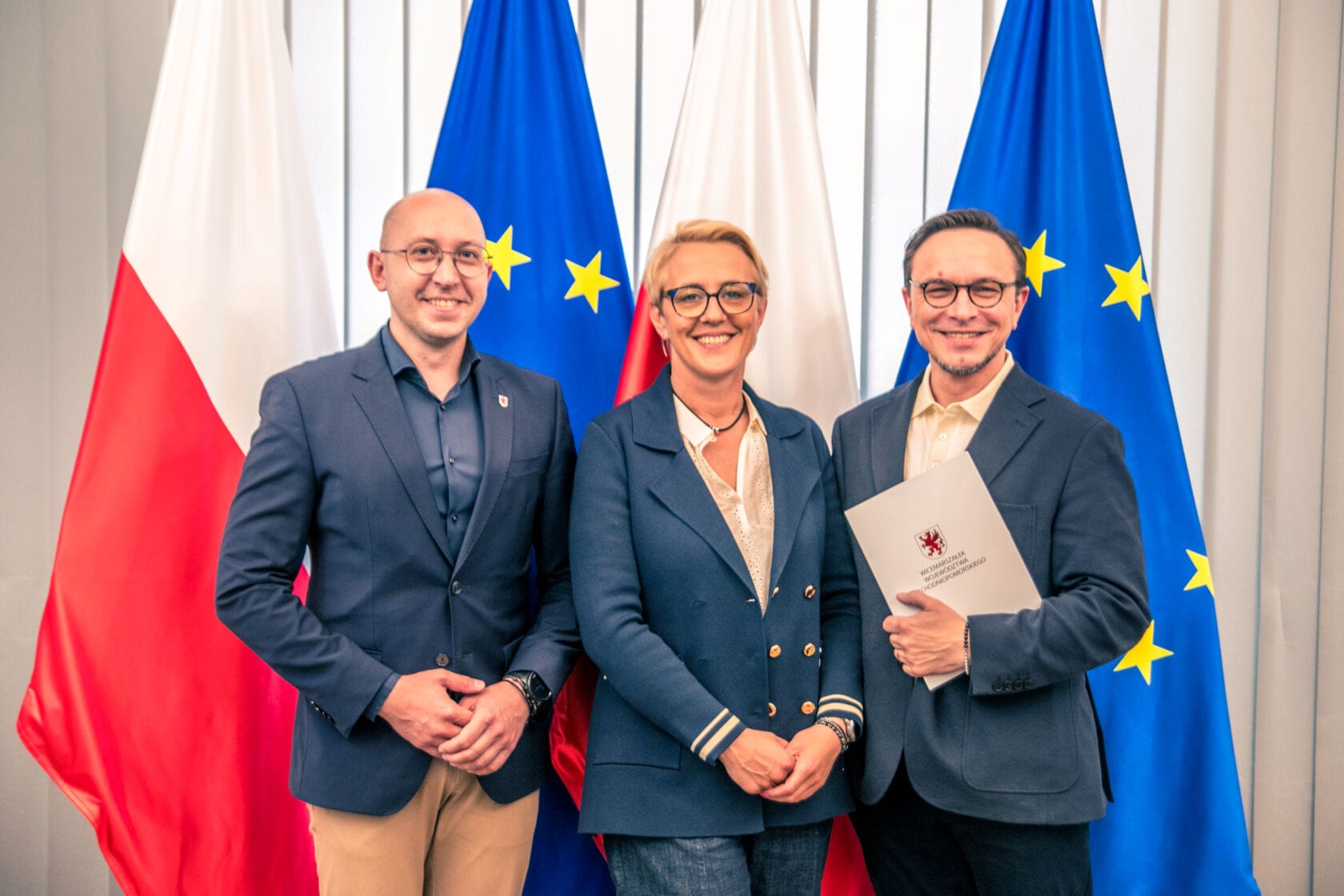 Trzy osoby uśmiechają się, trzymając dokument, flagi Polski i UE