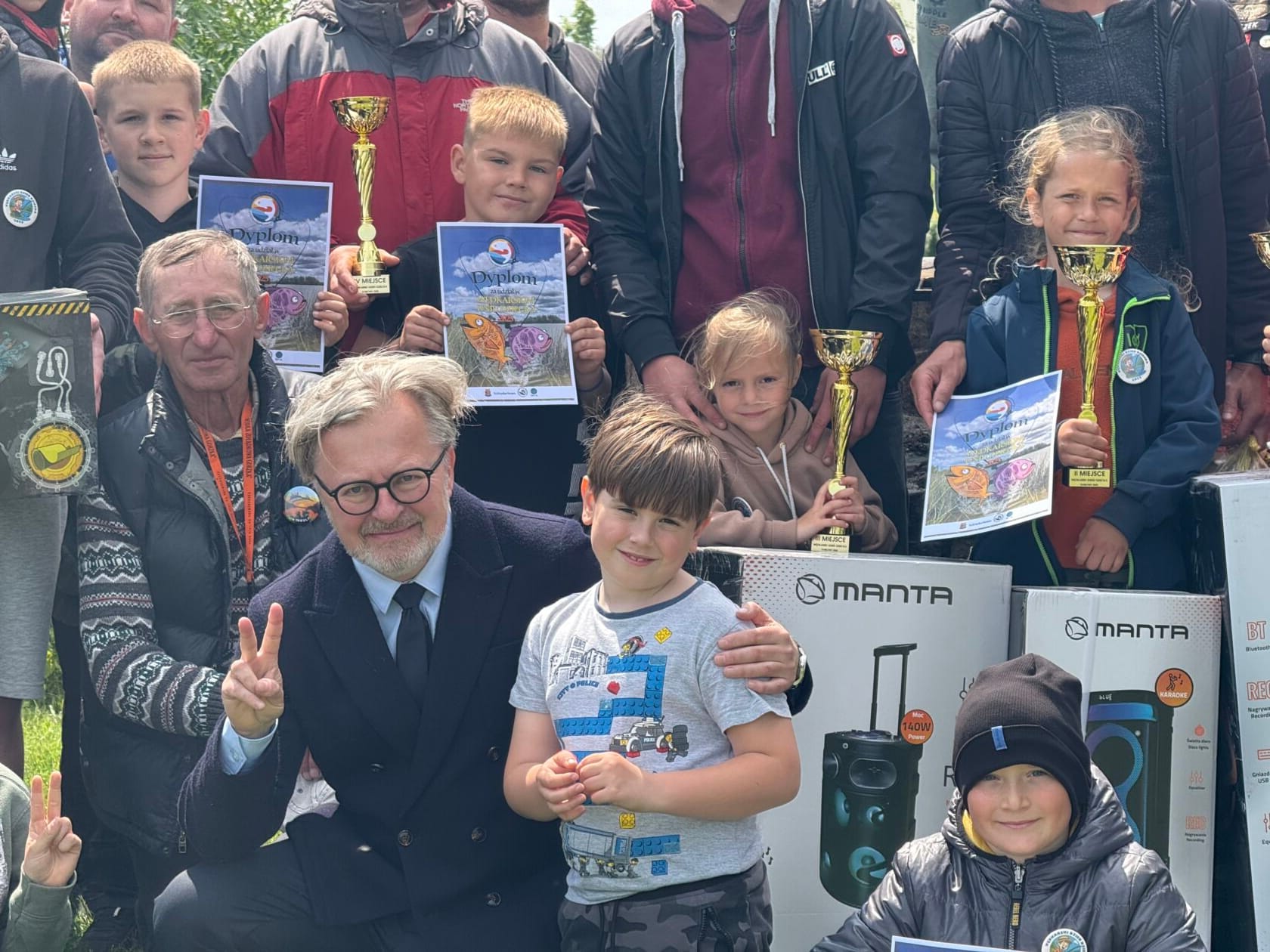 Grupa osób z pucharami i dyplomami celebruje na zewnątrz.