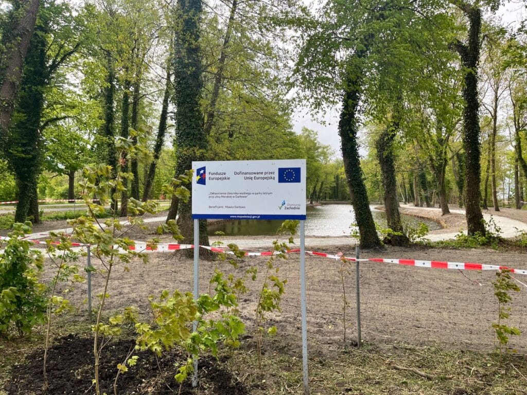 Park przy stawie z tablicą informacyjną i taśmą ostrzegawczą